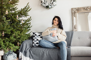 Happy young Caucasian woman with curly hair sitting on grey sofa near the Christmas tree, holding a pillow and looking to the camera with a lovely smile. Christmas and New Year concept.