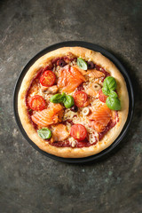 Traditional pizza with smoked salmon, cheese, tomatoes and basil served on black plate over old dark metal background. Top view with space. Rustic style