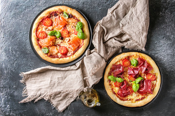 Two Traditional pizza with bresaola, smoked salmon, cheese, tomatoes and basil served on black plates with textile over black texture background. Top view with space.