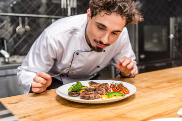 handsome chef standing with fork above fried vegetables with meat