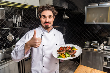 smiling chef showing thumb up and holding cooked vegetables with meat