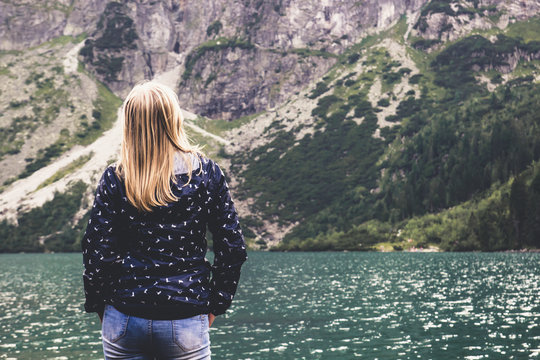 A young blonde girl stands back and looks at blue mountain lake and the mountains