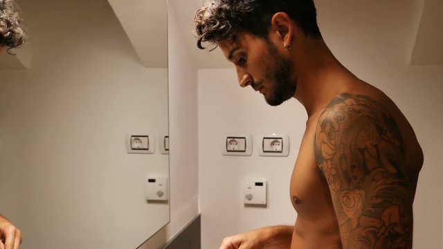Attractive shirtless young man brushing teeth with toothbrush in his bathroom, looking at himself in mirror