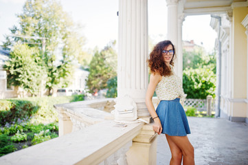 Curly stylish girl wear on blue jeans skirt, blouse and glasses posed near old vintage house.