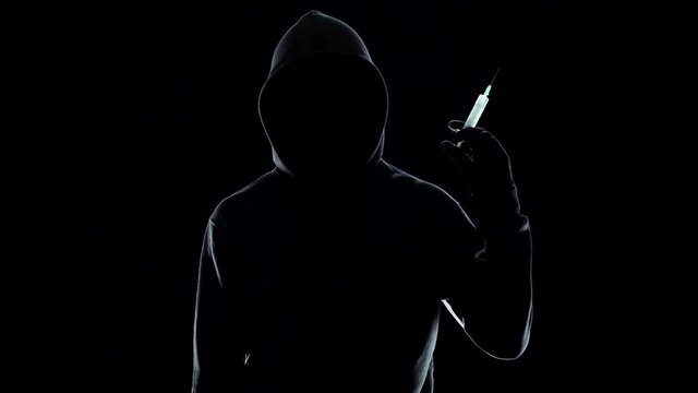 Unrecognizable hooded male holding syringe on black background, drug concept