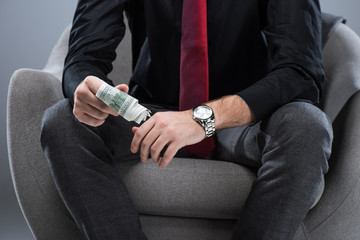 Midsection of businessman holding money in hands, while sitting on armchair, isolated on gray