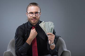 Successful smiling man with money in hand sitting in armchair while straightening tie, isolated on gray