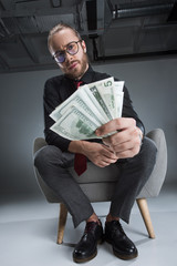 Serious businessman in glasses sitting in armchair with money in hands and looking at camera