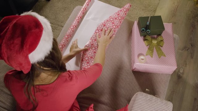 A woman in a Christmas hat measures wrapping paper for a gift box on a couch - top view