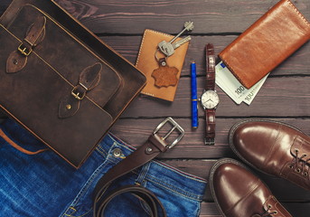 Top view, men's fashion personal belongings and accessories with space on a dark wooden background. Leather bag, shoes, watch, jeans, belt.
