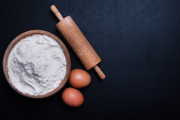 White flour in wooden bowl, eggs and rolling pin on black background with space for text