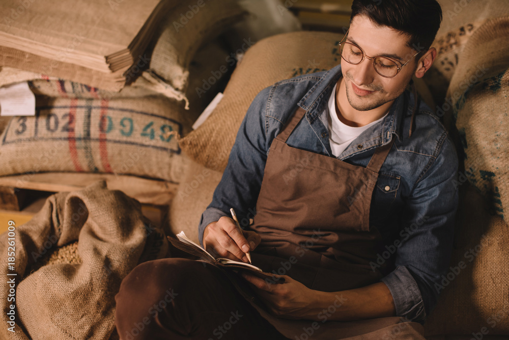 Wall mural portrait of smiling coffee roaster in eyeglasses and apron making notes in notebook