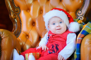 beautiful toddler in santa claus cap sits on chair smiling and looks at camera.