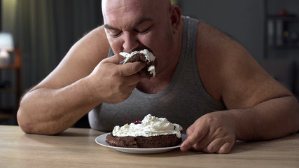 Messy obese man greedily eating cake with whipped cream, addiction to sweets