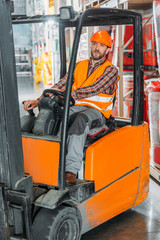 Fototapeta na wymiar male worker in safety vest and helmet sitting in forklift machine in storage