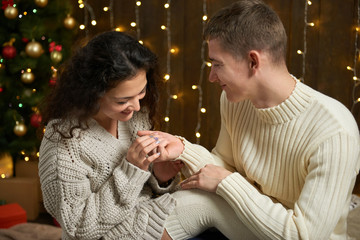 the man gives the girl an engagement ring, couple in christmas lights and decoration, dressed in white, fir tree on dark wooden background, winter holiday concept