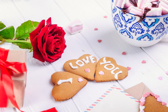 Homemade cookies in shape of heart with I Love you words on the white wooden table with greeting card, flower, gift box and cup of cacao. Gift for lover on Valentine's day. Selective focus.