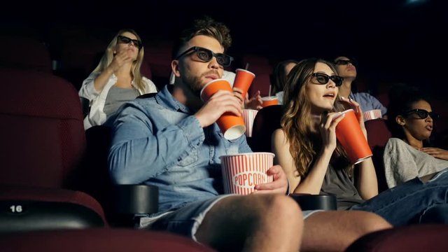 Young focused men and women hipsters in 3d glasses watching movie and eating popcorn in cinema