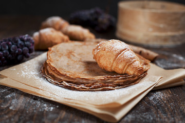 freshly baked sweet pancakes stacked on brown baking paper, croissants and grapes on a dark brown wooden table on a dark gray background