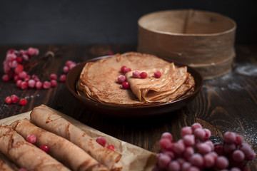 Fototapeta premium sweet fresh pancakes with a pile and pancakes on baking paper, rolled with tubules, viburnum berries on a dark wooden table on a dark gray background