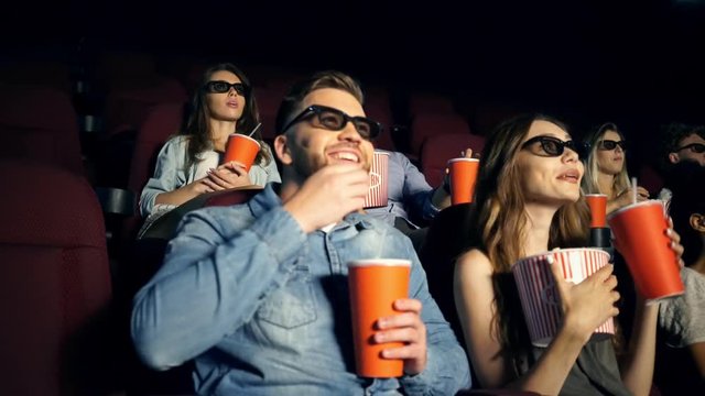 Cheerful young lovers in 3d glasses talking while watching movie and eating popcorn in cinema