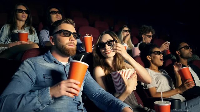 Cheerful lovers hipsters talking and smiling while watching film and holding cola and popcorn on cinema
