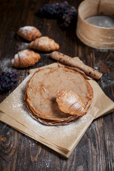 freshly baked sweet pancakes stacked on brown baking paper, croissants and grapes on a dark brown wooden table on a dark gray background