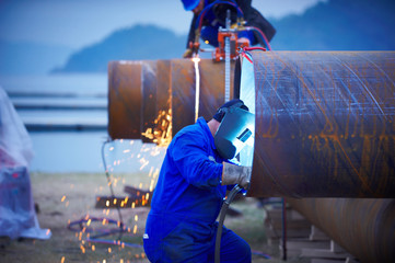 Industrial electrode welder with face shield and blue overall welding a steel pipe in workshop.