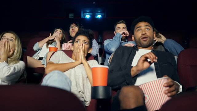 Shocked multiethnic young people watching film with popcorn in cinema and frightened