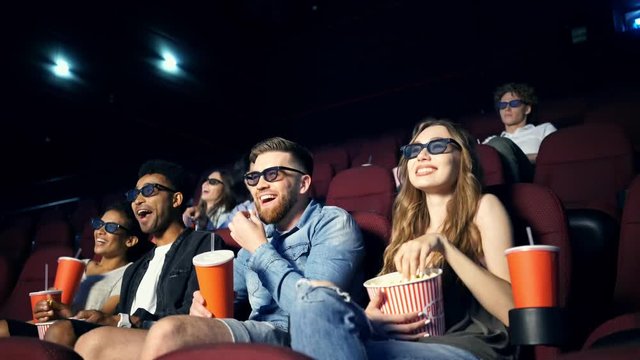 Laughing multiethnic young friends wearing 3d glasses eating popcorn and watching movie in cinema