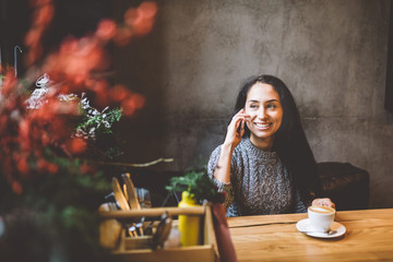 beautiful young brunette girl talking on mobile phone at wooden table near window and drinking coffee in cafe decorated with Christmas decor. Dressed in a gray knitted wool sweater