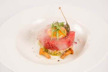 Smoked steak tartare served with egg yolk,  onion, pickle,  caper, mushroom and rocket salad on a white background