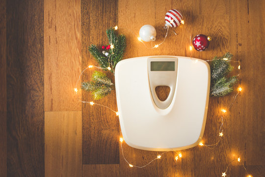 Top View Of A White Digital Weight Scale On Wooden Background Surrounded With Christmas Lights And Decoration And Snowy Pine Tree Branches, Weight Gain During Holidays Concept, Vintage Toned