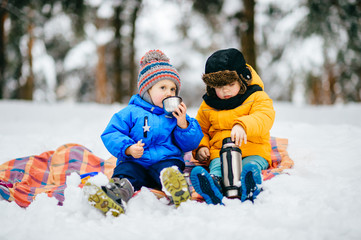 Fototapeta na wymiar Funny children winter party in snowy forest. Kids male friends rest outdoor at nature. Young boys sharing and drinking tea from thermos. Hot drinks and beverage in cold weather. Warming in frost day.