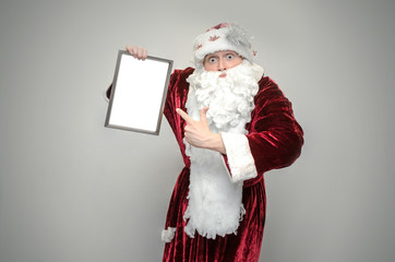 Santa Claus hold in hands a blank paper page in photo frame with copy space isolated on gray background. Christmas congratulatory letter.
