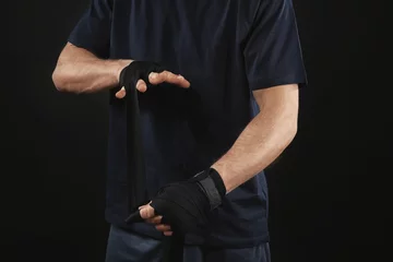 Abwaschbare Fototapete Kampfkunst Male boxer applying wrist wraps on black background, closeup