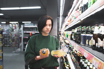 Portrait of a girl who chooses beer in a supermarket. The girl looks at the alcohol in his hands....