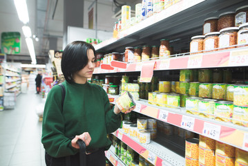 The girl chooses canned produce from supermarket shelves. The girl is shopping at the supermarket.