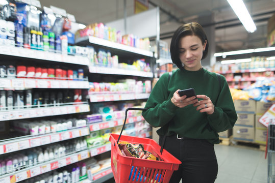 A Positive Girl Uses A Phone In A Supermarket. A Girl Shopping In A Supermarket With A Phone In Her Hands. Focus On Your Phone