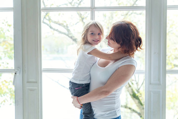 Young mother holding her child. Woman and little girl relax in a white bedroom near the windiow. Happy family at home. Young mother playing with her daughter