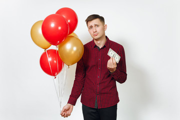 Handsome caucasian sad upset young y man in red plaid shirt with yellow golden balloons, one dollar banknote celebrating birthday, on white background isolated for advertisement. Holiday, pure concept