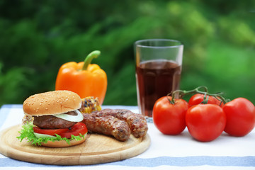 Wooden board with delicious hamburger and grilled sausages on table outdoors