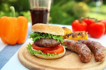 Wooden board with delicious hamburger and grilled sausages on table outdoors