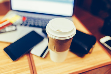 White paper Cup with burning coffee on the background of laptop and smartphone. Scattered Bank cards and a black purse. Work on the go. Toned photo.