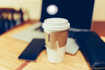 White paper Cup with burning coffee on the background of laptop and smartphone to Work on the go. Toned photo.