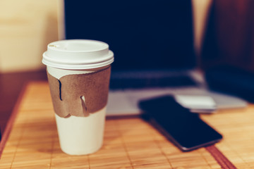 White paper Cup with burning coffee on the background of laptop and smartphone to Work on the go. Toned photo.