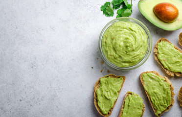 Traditional mexican homemade guacamole sauce in a glass bowl and sliced bread on a light gray stone background. Top view, copy space, horizontal - obrazy, fototapety, plakaty
