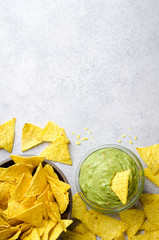 Traditional mexican homemade guacamole sauce in a glass bowl and a bowl with tortilla chips on a light stone background. Party food concept.  Top view, copy space, vertical