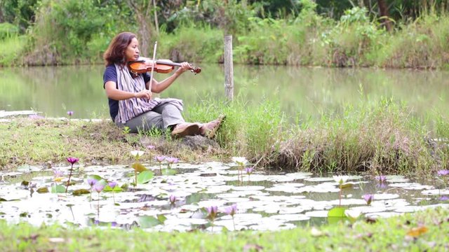 an asian woman with black hair, jean and scarf plays violin near the lake and lotus pool