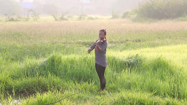 an asian woman playing violin on grass field in the morning